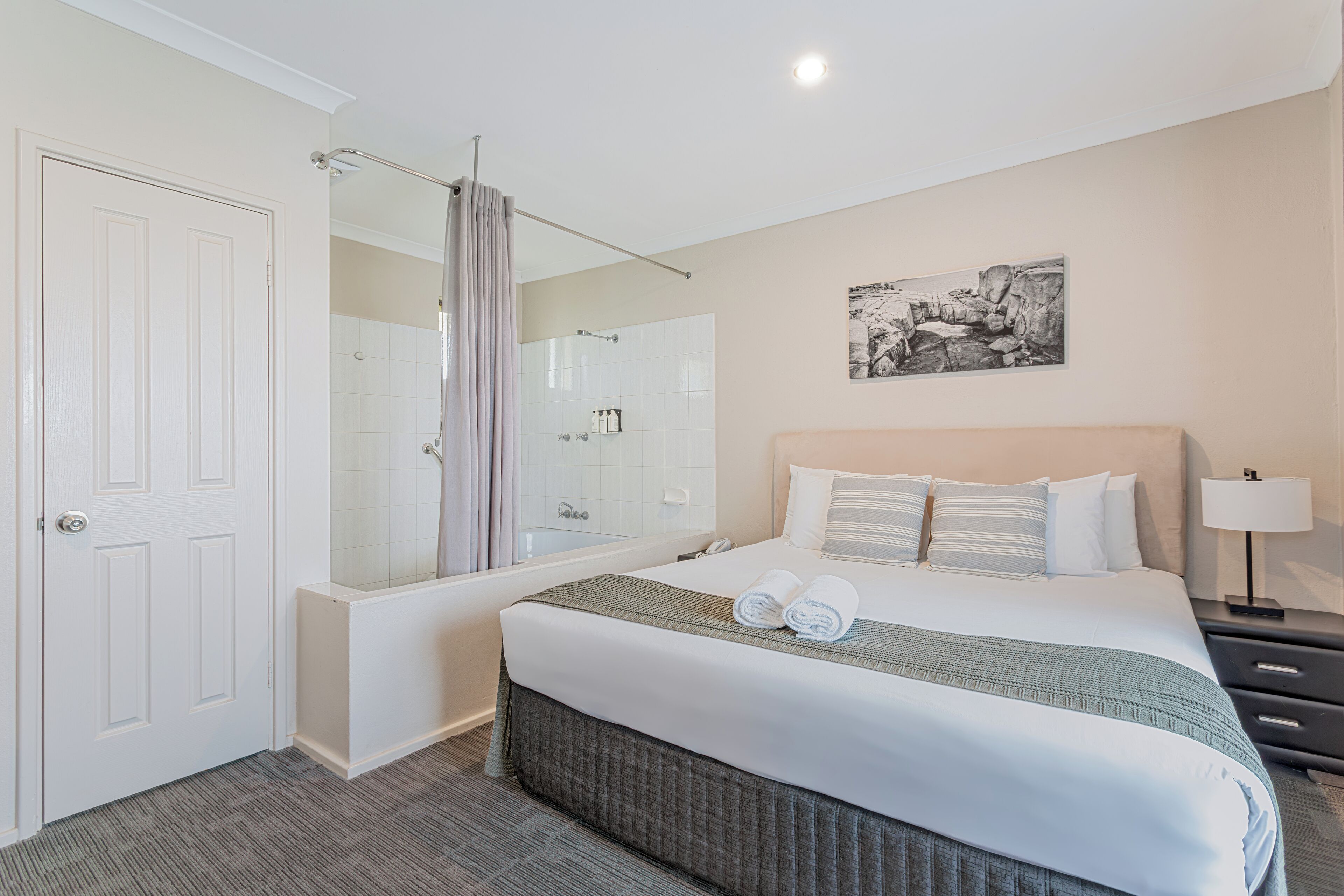 A hotel room with a large bed, a shower area, a door, and a nightstand with a lamp.