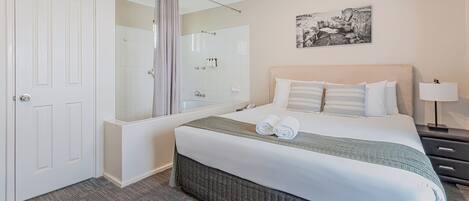 Una habitación de hotel con una cama grande, zona de ducha, una puerta y una mesita de noche con lámpara.