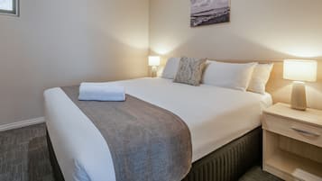 Una cama bien hecha con una manta gris, dos lámparas de noche, una mesita de madera y un cajón pequeño.