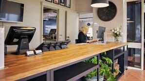 A reception desk with a wooden top, a black desk lamp, and a digital display. There is a clock on the wall and a plant in the background. - Mandala Ace Albany Hotel (Centennial Park)
