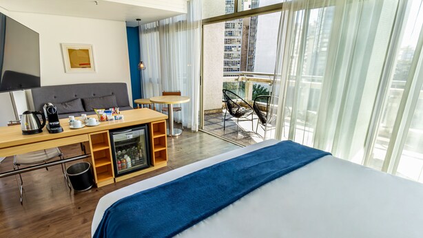 A hotel room with a bed, a desk with a coffee maker, a mini-fridge, and a balcony with chairs. - Hotel Nacional Inn Jaragua São Paulo (São Paulo)