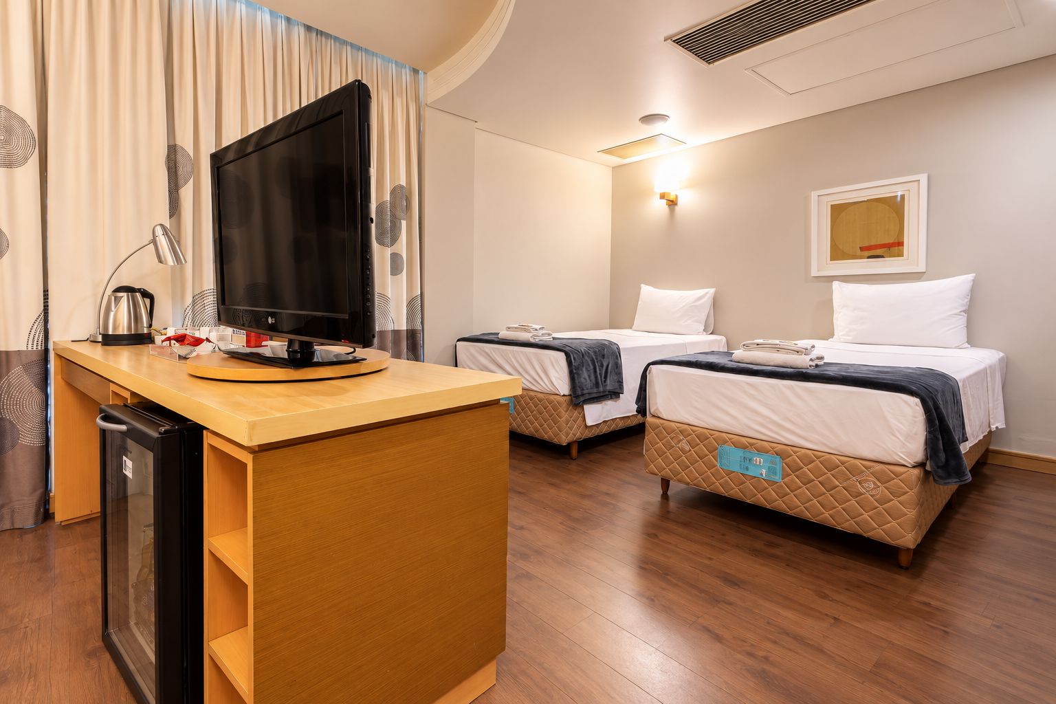 Quarto de hotel com escrivaninha de madeira, televisão de tela plana, duas camas com colchas, pequeno frigorífico e luminária fixada na parede.