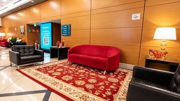 A hotel lobby with a red sofa, black armchairs, a patterned carpet, and a digital information board.