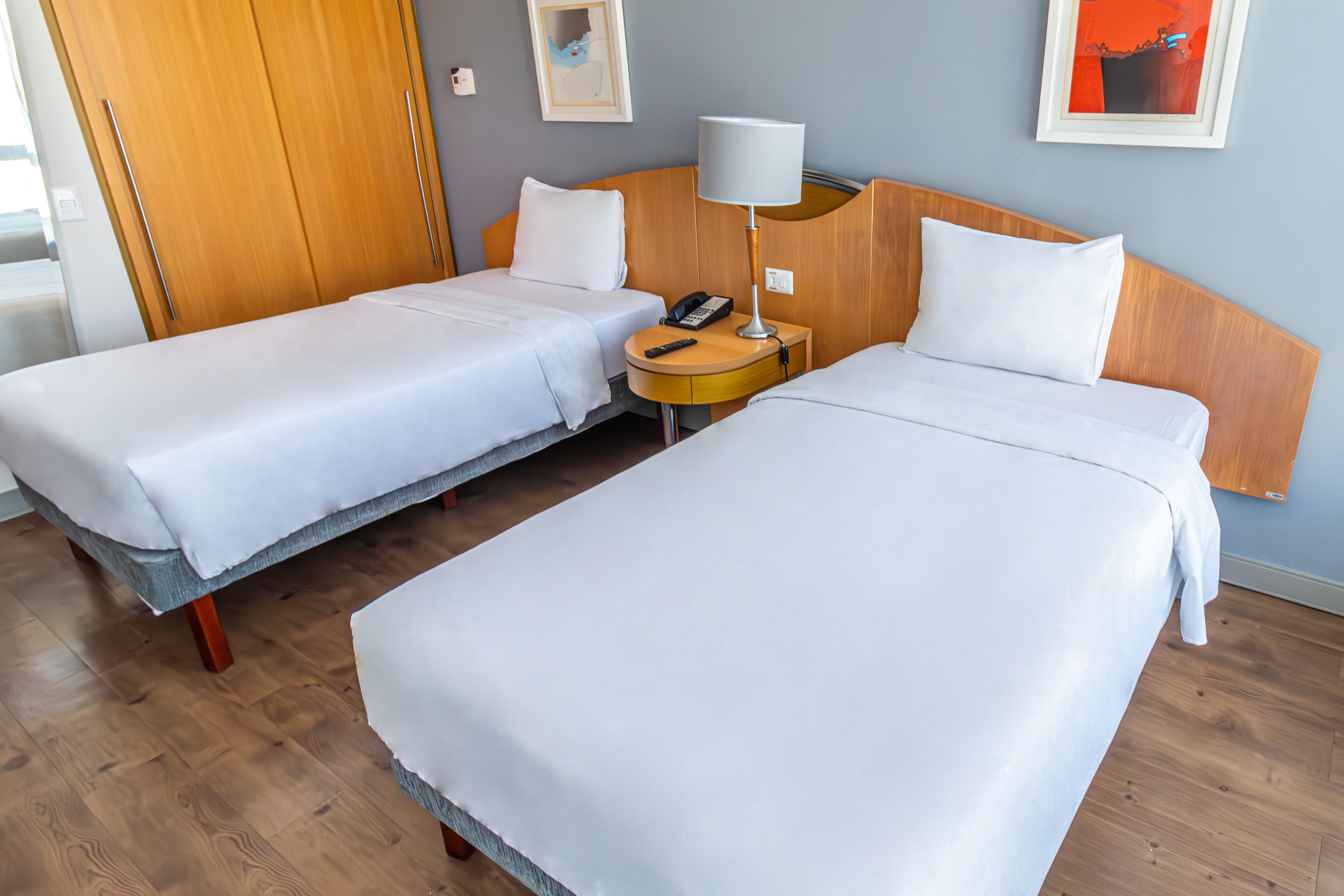 A hotel room with two single beds, a wooden wardrobe, a bedside table with a lamp, and a framed picture on the wall.