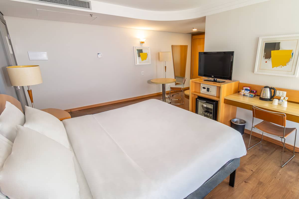 Hotellihuone, jossa on suuri sänky, työpöytä tietokoneella, televisio ja pieni jääkaappi.