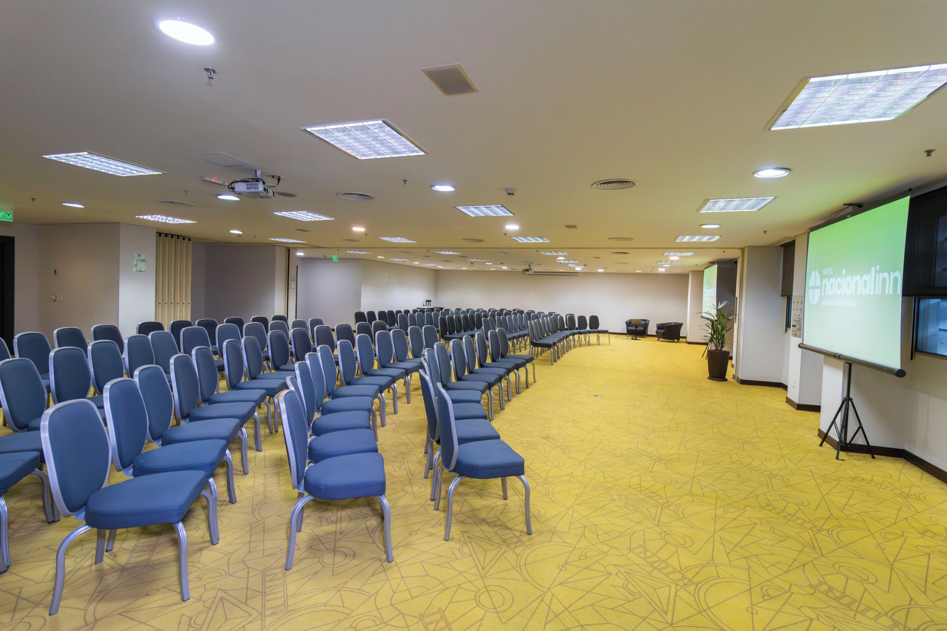 A conference room with rows of blue chairs, a projector screen, and a yellow patterned carpet.