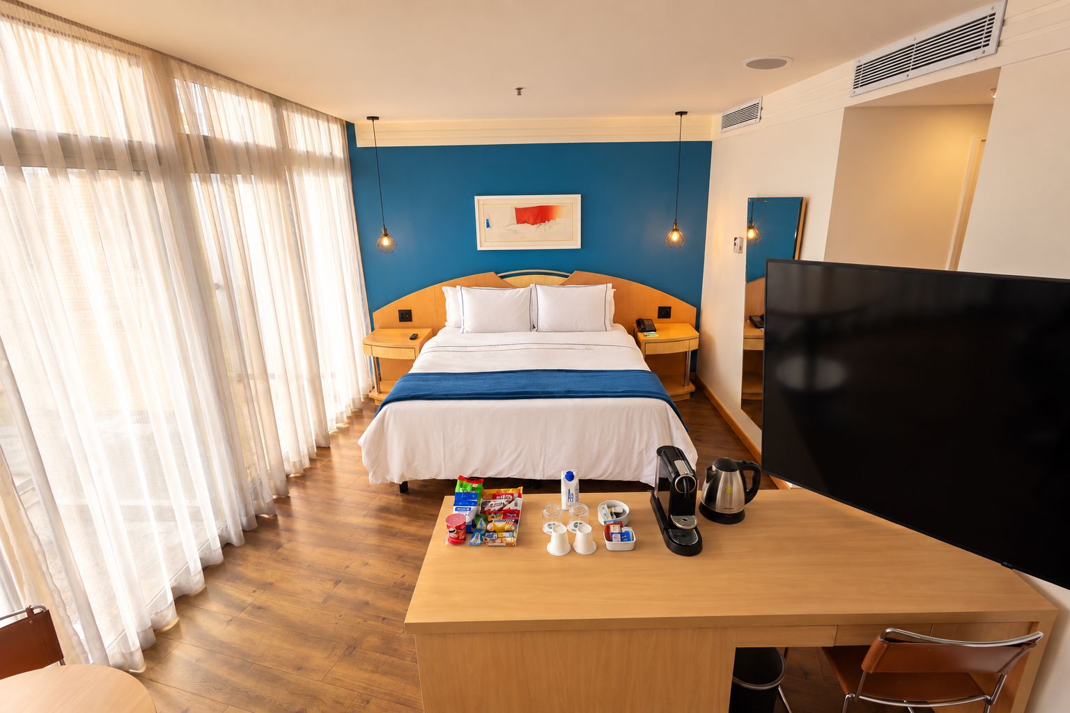 Habitación de hotel con cama, mesitas de noche, escritorio con silla, televisor y ventana con cortinas.