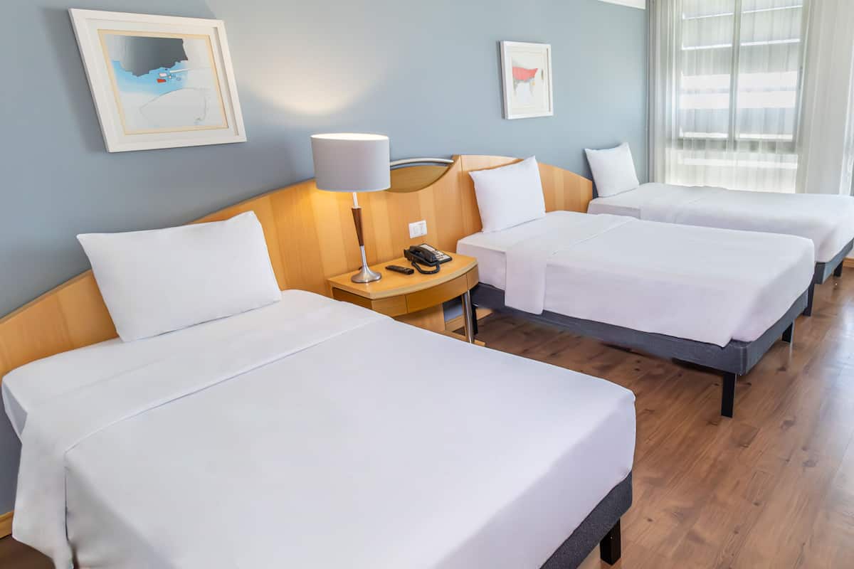 Hotellihuone, jossa on kolme erillistä sänkyä, puinen pääty, yöpöytä lampun kanssa, puhelin ja seinillä tauluja.