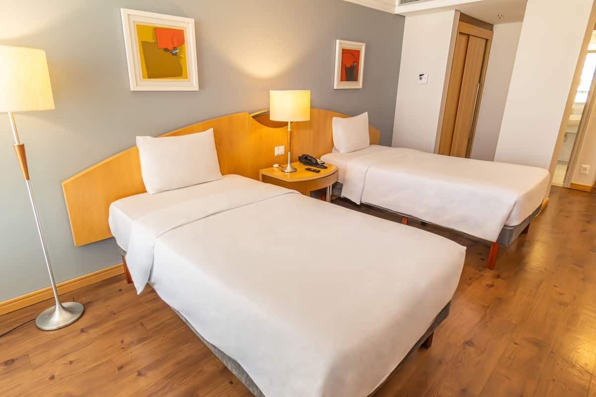 Hotellihuone, jossa on kaksi sänkyä, yöpöytä lampun kanssa ja taideteoksia seinillä.