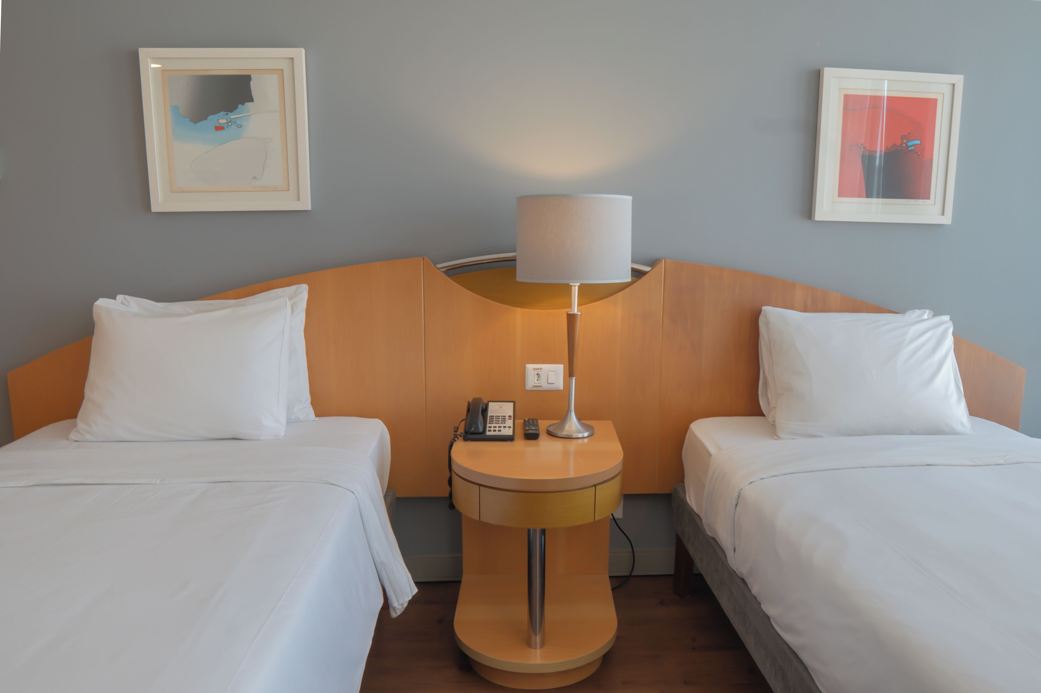 Quarto de hotel com duas camas de solteiro, uma mesa de cabeceira com abajur, telefone e duas imagens emolduradas na parede.