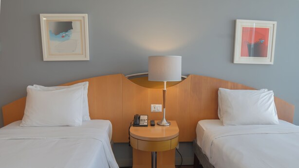 Habitación de hotel con dos camas individuales, una mesita de noche con lámpara, teléfono y dos cuadros en la pared.