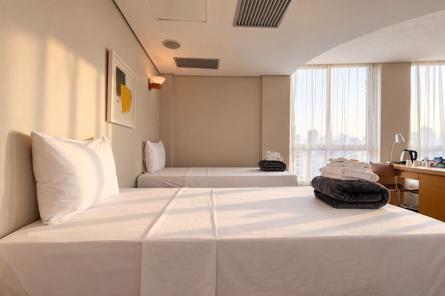 A hotel room with two beds, a desk, a chair, a window with curtains, and a city view.