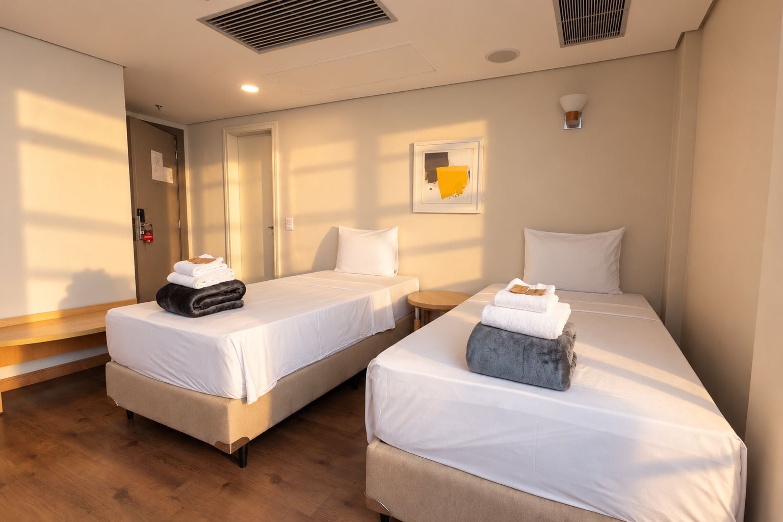 Habitación de hotel con dos camas, piso de madera, una mesita redonda y una lámpara de pared.
