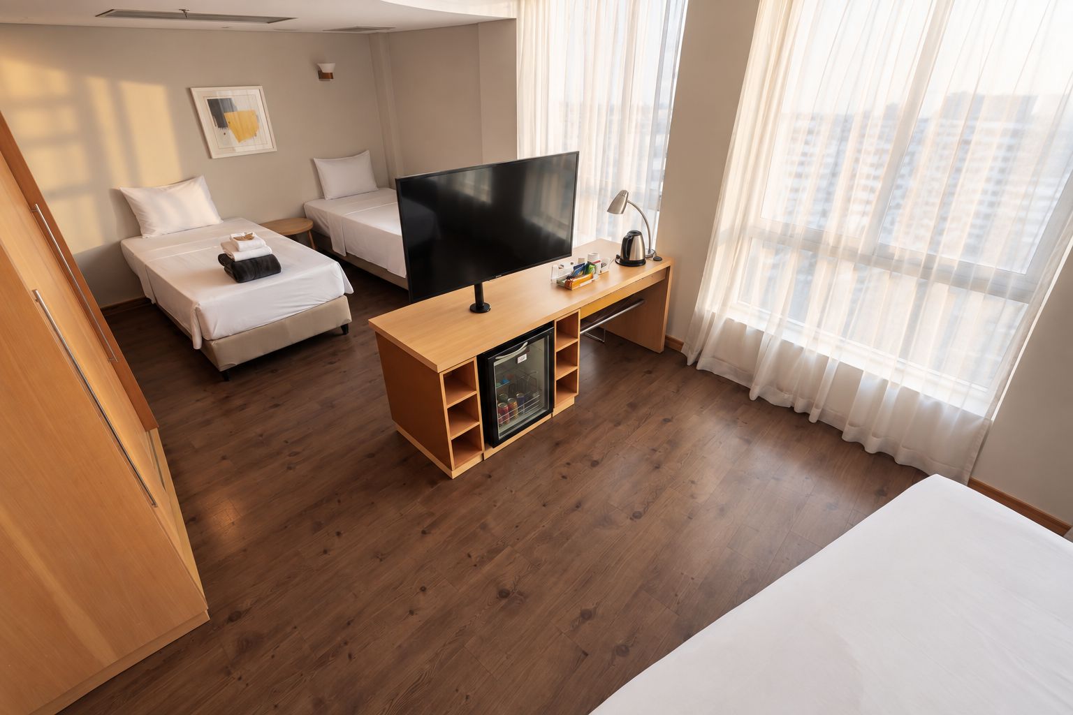 Quarto de hotel com cama, televisão, escrivaninha com abajur e janela com cortinas.