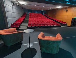 Un auditorio con hileras de asientos rojos, un escenario y dos sillones verdes.