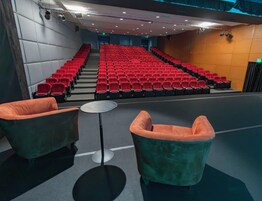 Un auditorium con file di posti a sedere rossi, un palco e due poltrone verdi.