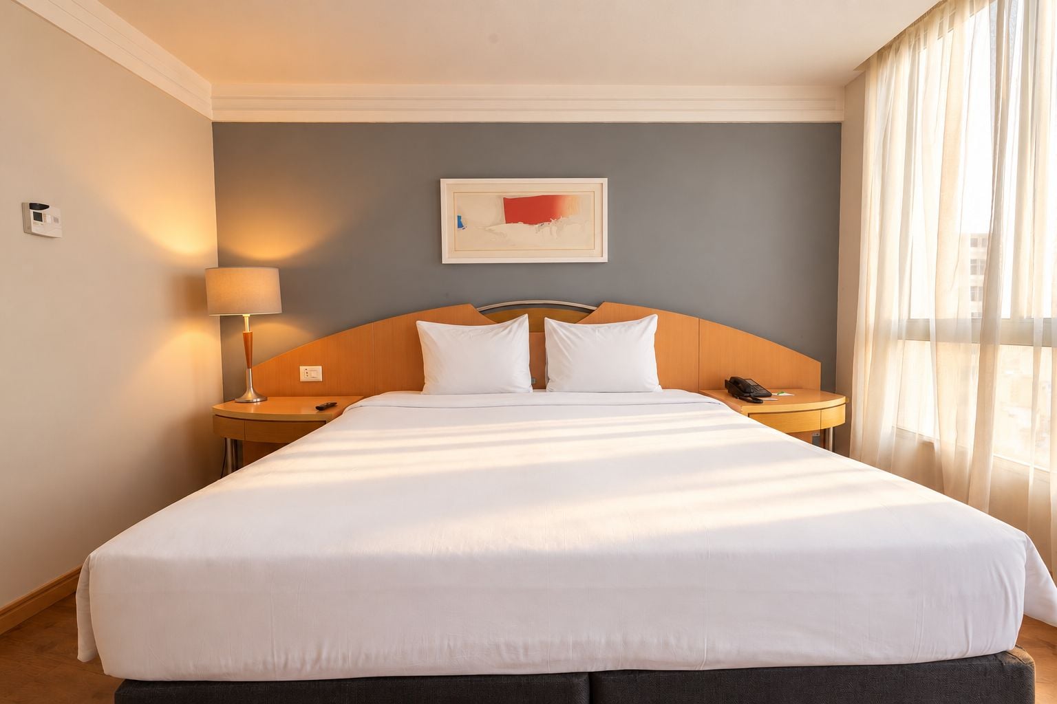 Quarto de hotel com uma cama grande, mesa de cabeceira com abajur, telefone e janela com cortinas.