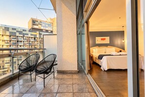 Un balcon avec deux chaises, un lit avec une literie blanche et une vue sur la ville.