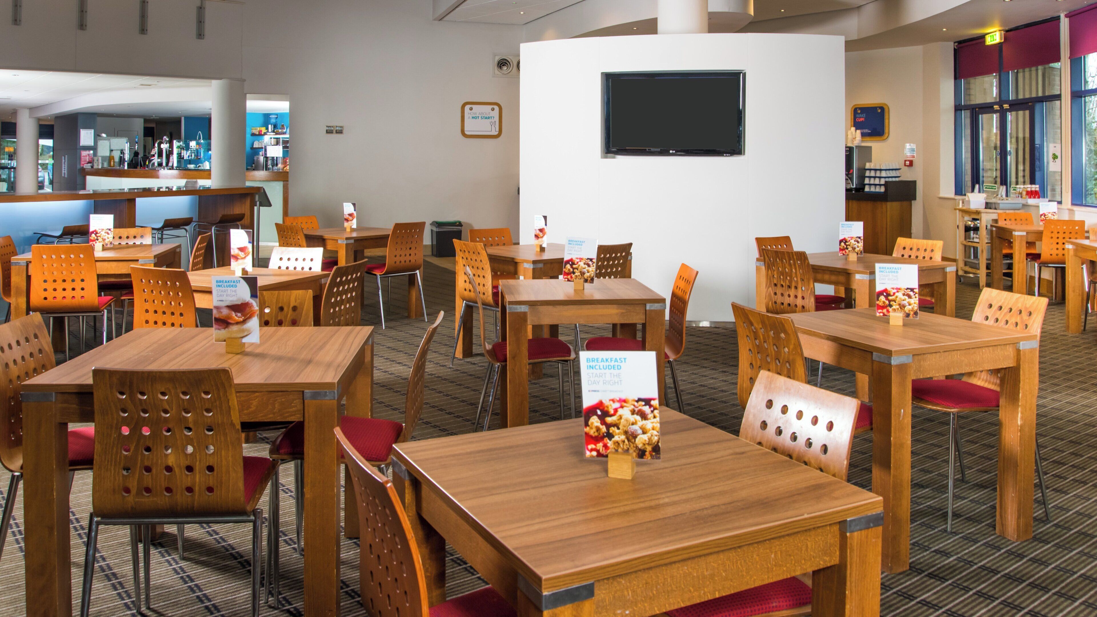 A hotel dining area with wooden tables and chairs, breakfast included signs, and a TV mounted on the wall.