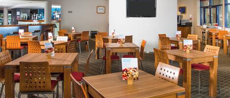 A hotel dining area with wooden tables and chairs, breakfast included signs, and a TV mounted on the wall.