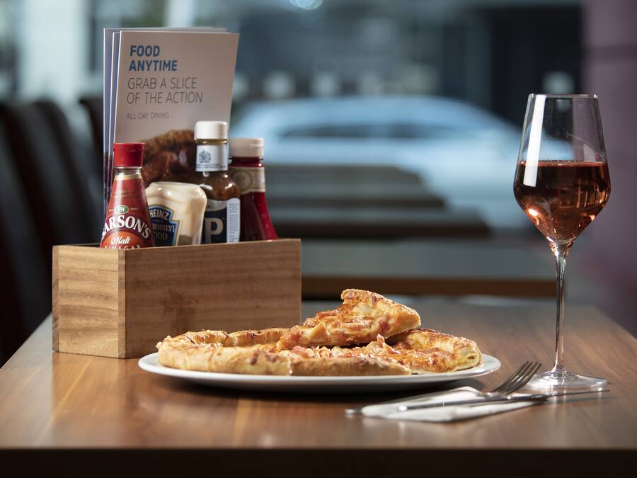 Una mesa de madera con una porción de pizza, una copa de vino tinto y condimentos en un caja de madera.