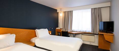 Et hotelværelse med to senge, et skrivebord, et fjernsyn og et vindue med gardiner.