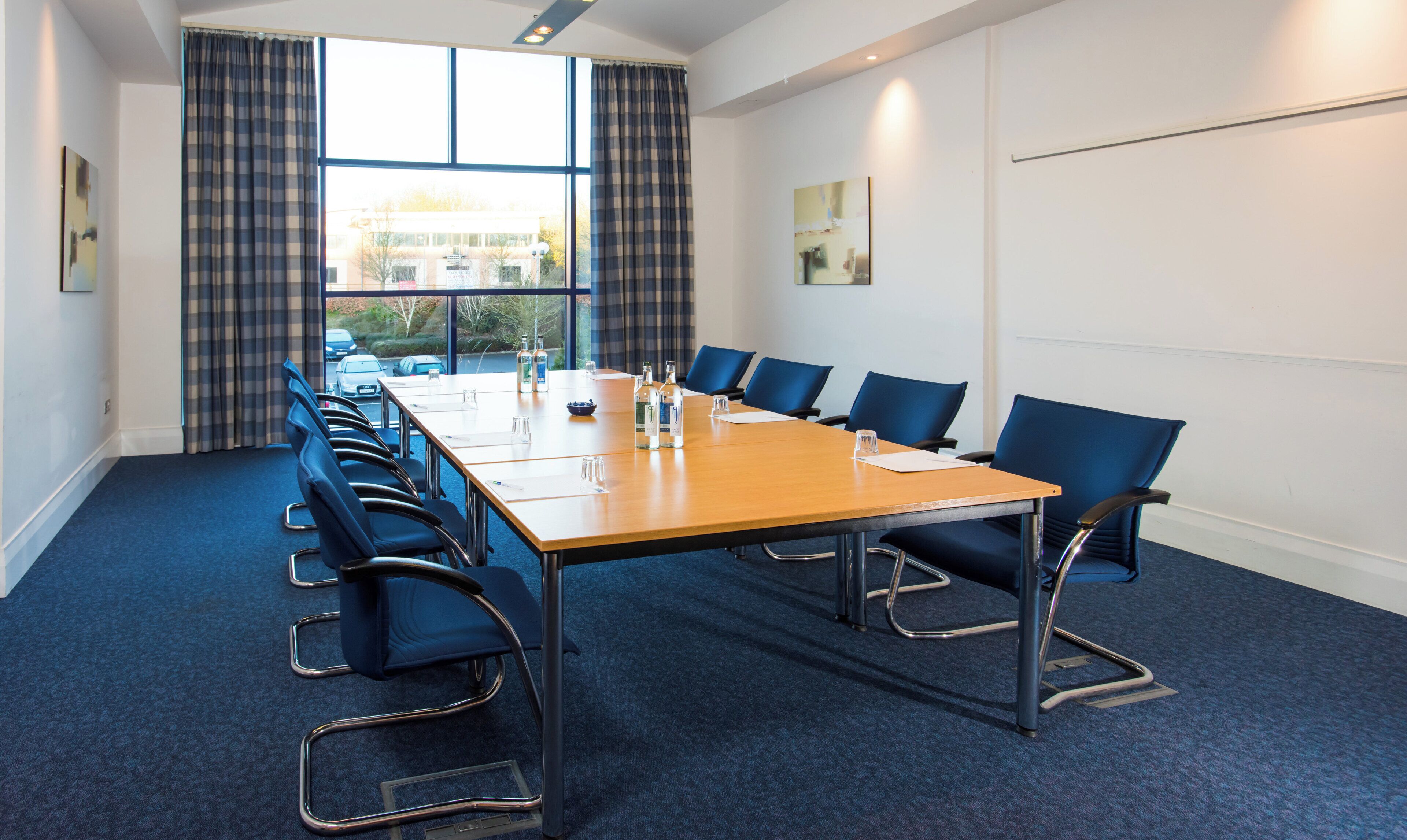A conference room with a long table, blue chairs, and a view of the outside through large windows.