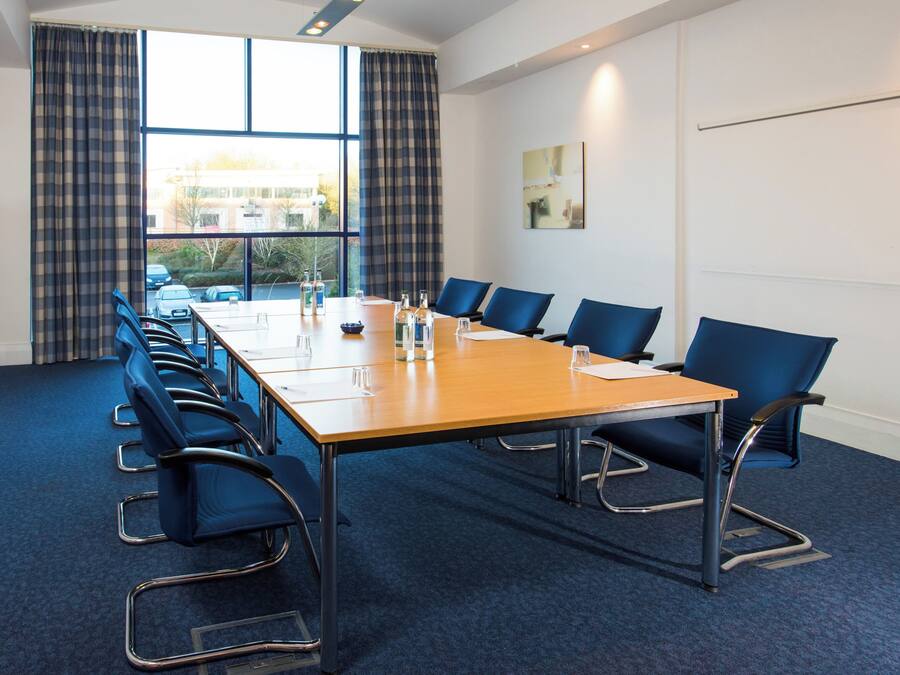 A conference room with a long table, blue chairs, and a view of the outside through large windows.