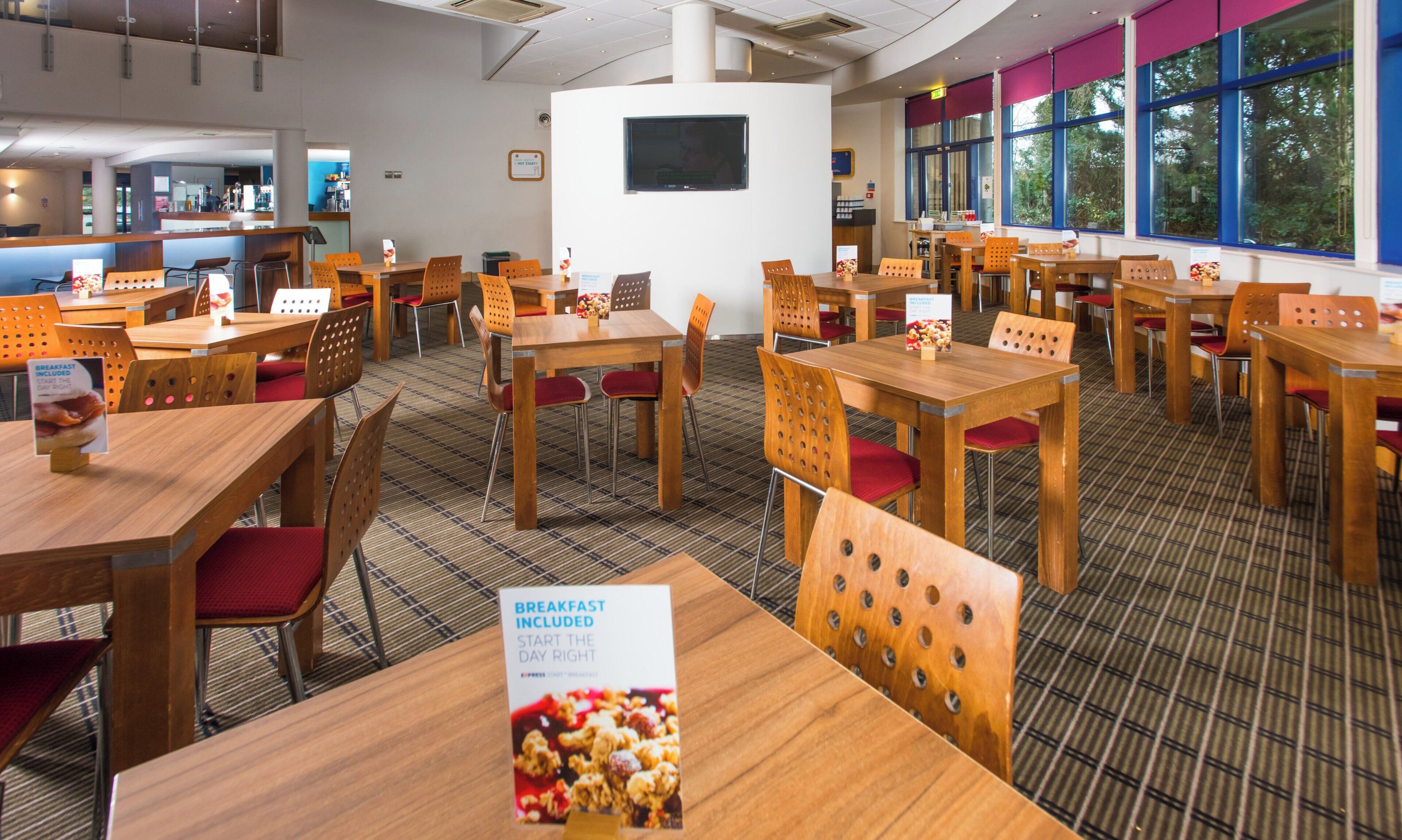 A restaurant with multiple tables and chairs, a sign indicating breakfast is included, and a large window with a view of trees.