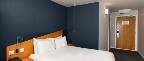 Une chambre d’hôtel avec un grand lit, une tête de lit en bois, une table de chevet avec une lampe et une porte surmontée d’un appareil de climatisation.