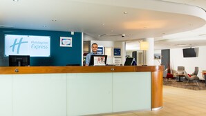 A Holiday Inn Express reception area with a wooden reception desk, a large screen displaying the hotel name, and a sign indicating the location of conference rooms.