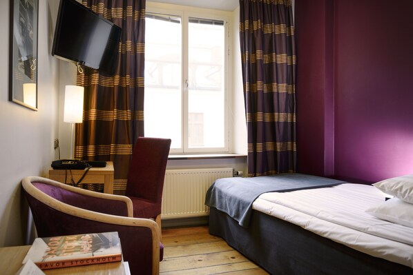 A hotel room with a bed, a chair, a desk with a book, a lamp, a TV, and curtains.