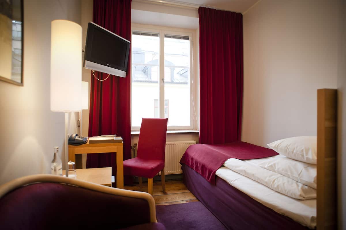 A hotel room with a bed, a desk with a chair, a TV, and a window with curtains.