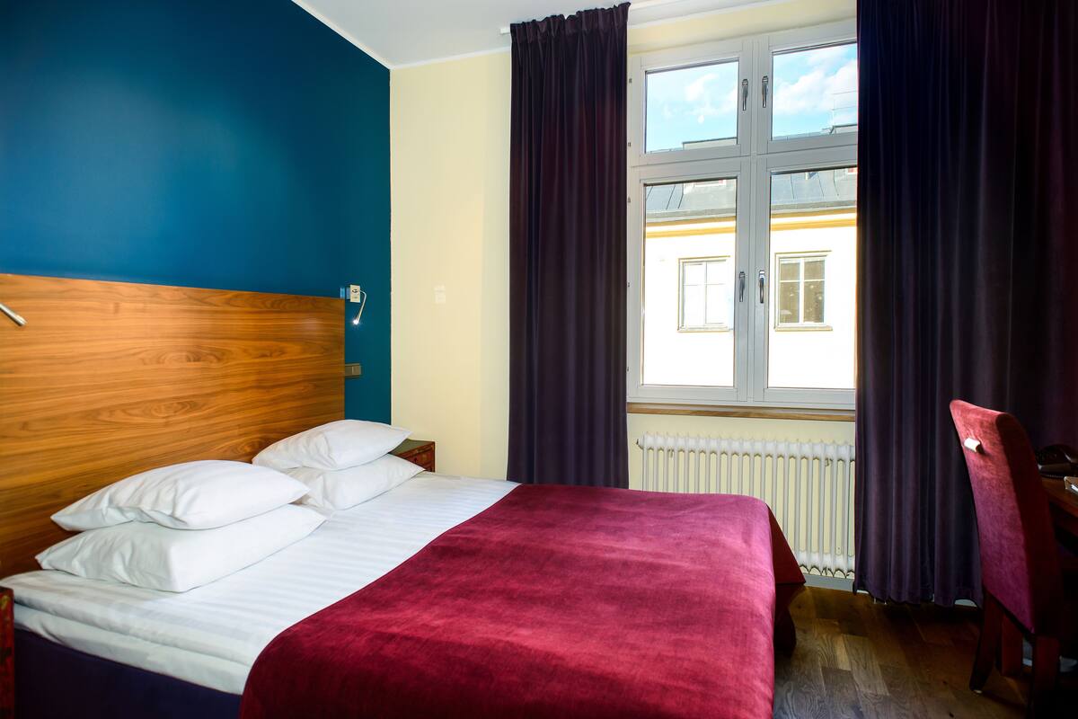 A hotel room with a wooden headboard, a bed with white linens and a red blanket, a window with curtains, and a chair.