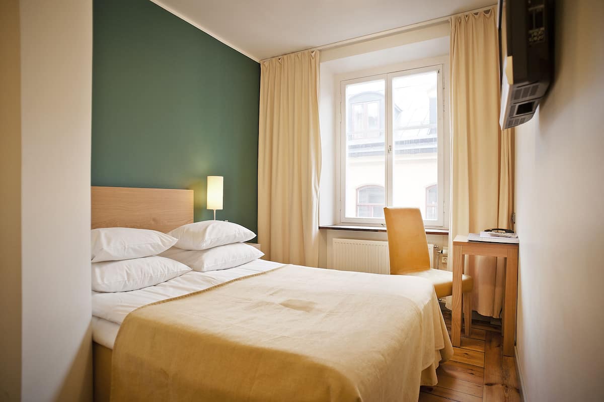 A hotel room with a bed, a desk, a chair, a window with curtains, and a wall-mounted TV.