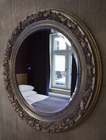 Um espelho com moldura decorada reflete um quarto com cama, janela e uma cadeira.