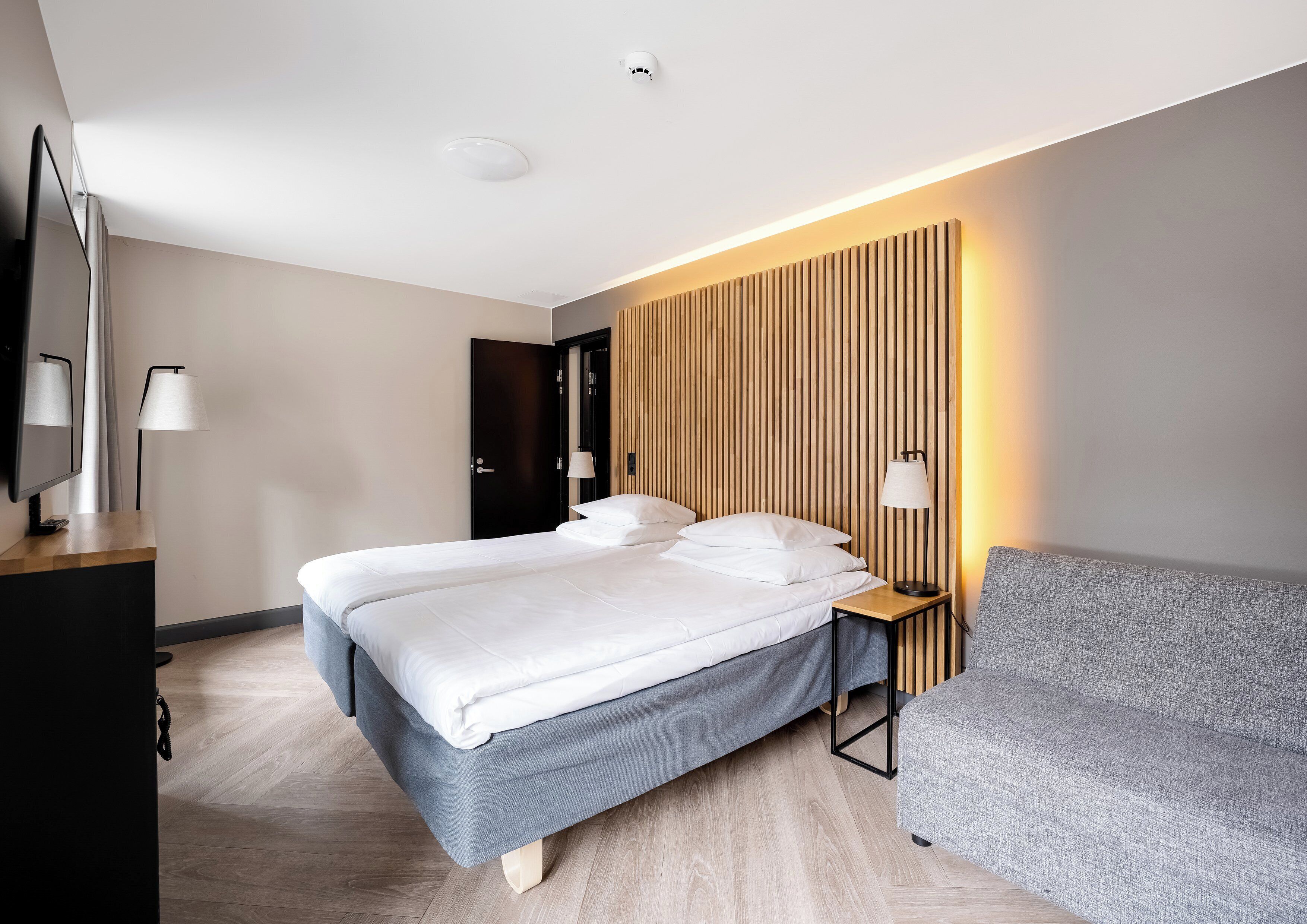 Una moderna camera d'albergo con un ampio letto, comodini, una TV a schermo piatto e una poltrona grigia.