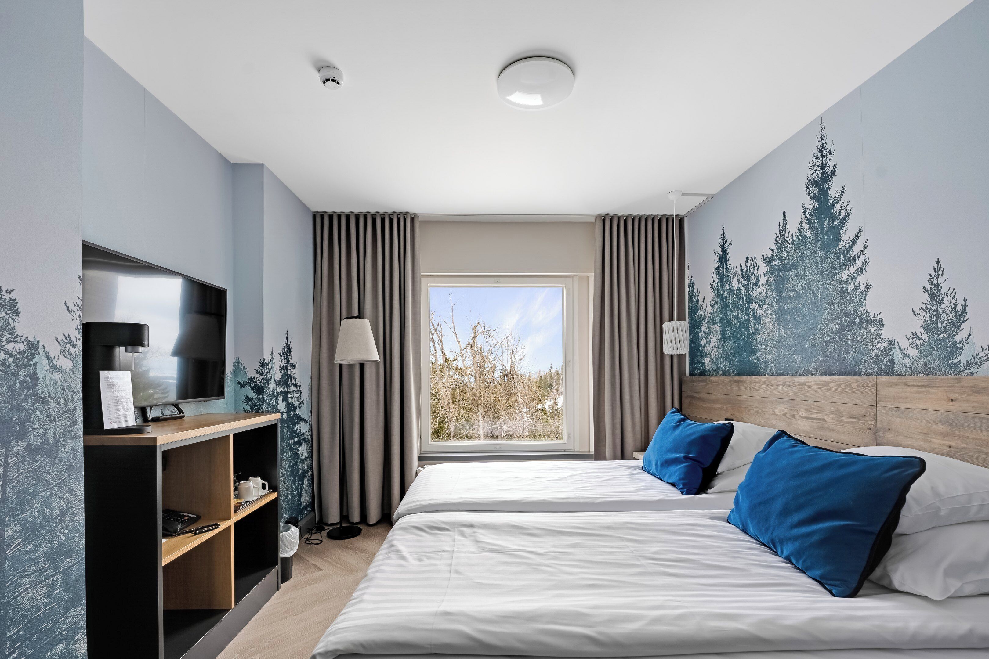 A modern hotel room with a large bed, a TV, a desk with a lamp, and a window with curtains.