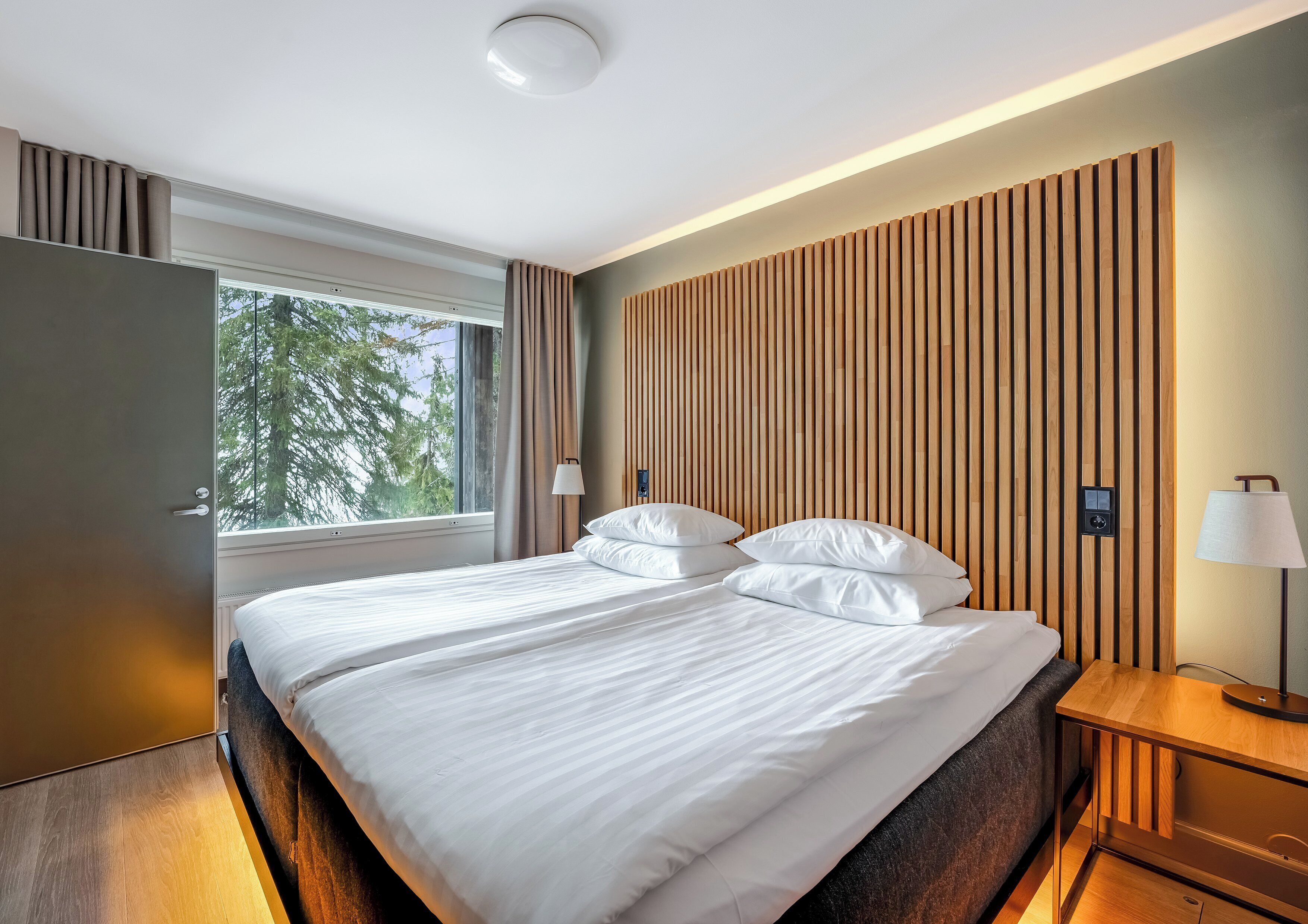 A modern hotel room with a large bed, wooden panel wall, and a window with curtains.