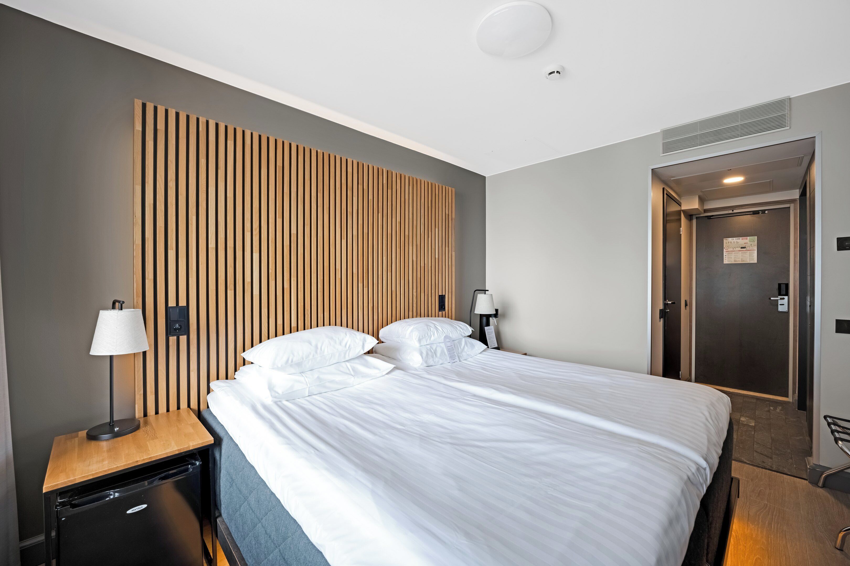 A modern hotel room with a large bed, a wooden headboard, a nightstand with a lamp, a small black refrigerator, and a door leading to a bathroom.