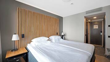 A modern hotel room with a large bed, a wooden headboard, a nightstand with a lamp, a small black refrigerator, and a door leading to a bathroom.