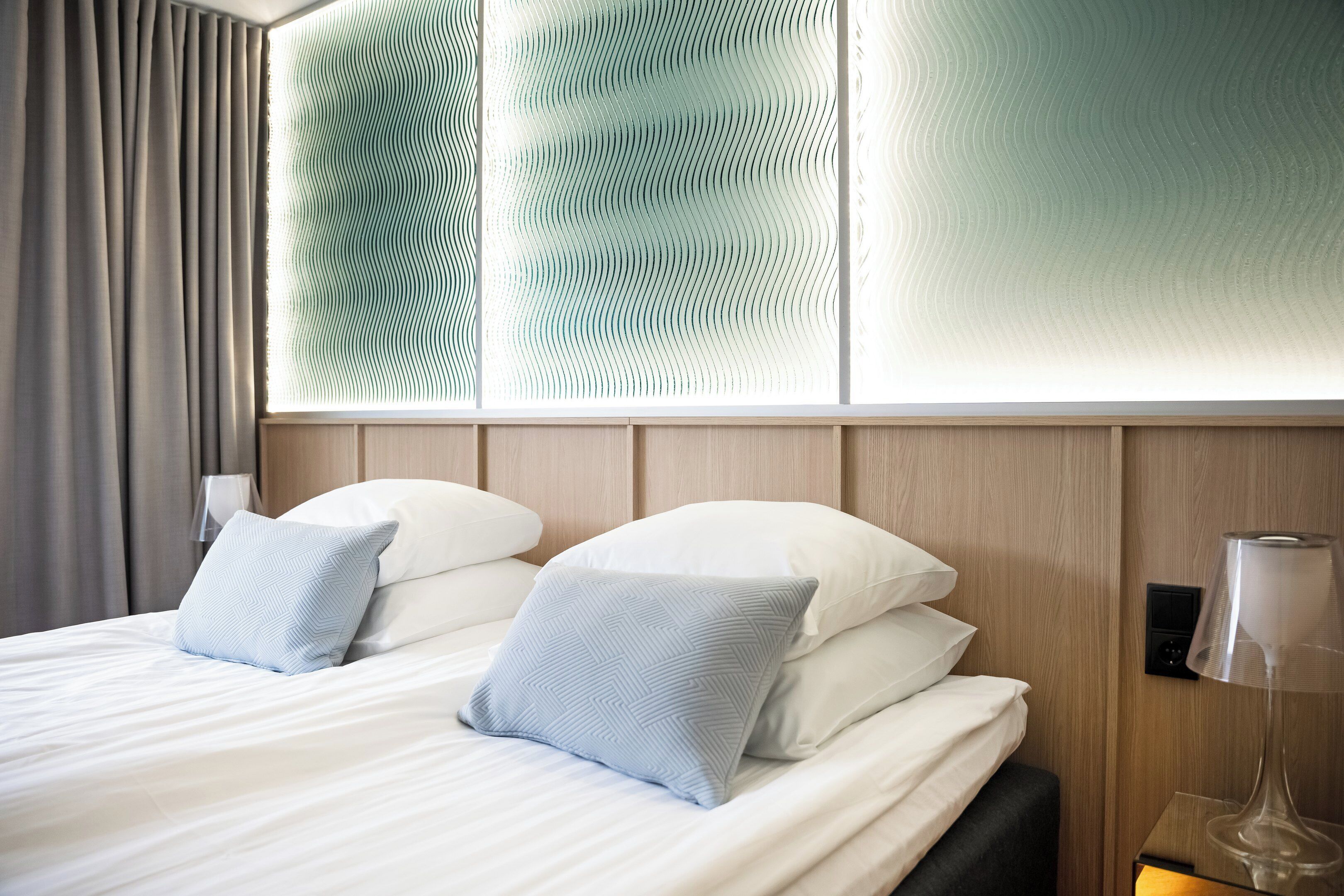 Hotelkamer met een bed, twee kussens, een nachtkastje met een lamp en een muur met een golvend patroon.