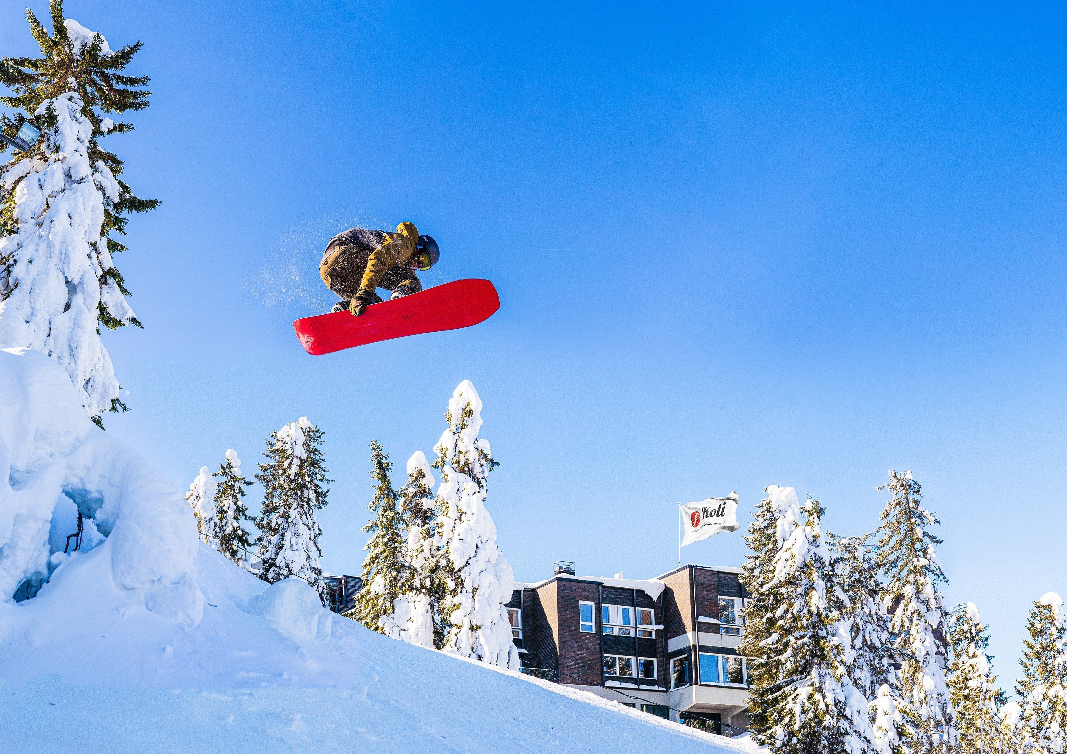 Un snowboardeur en plein vol au-dessus d’une pente enneigée, avec des bâtiments et des arbres en arrière-plan.
