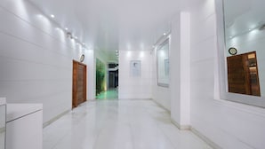 Un pasillo limpio y bien iluminado con paredes blancas y piso de baldosas, que conduce a una habitación con un ventanal grande y un reloj en la pared.