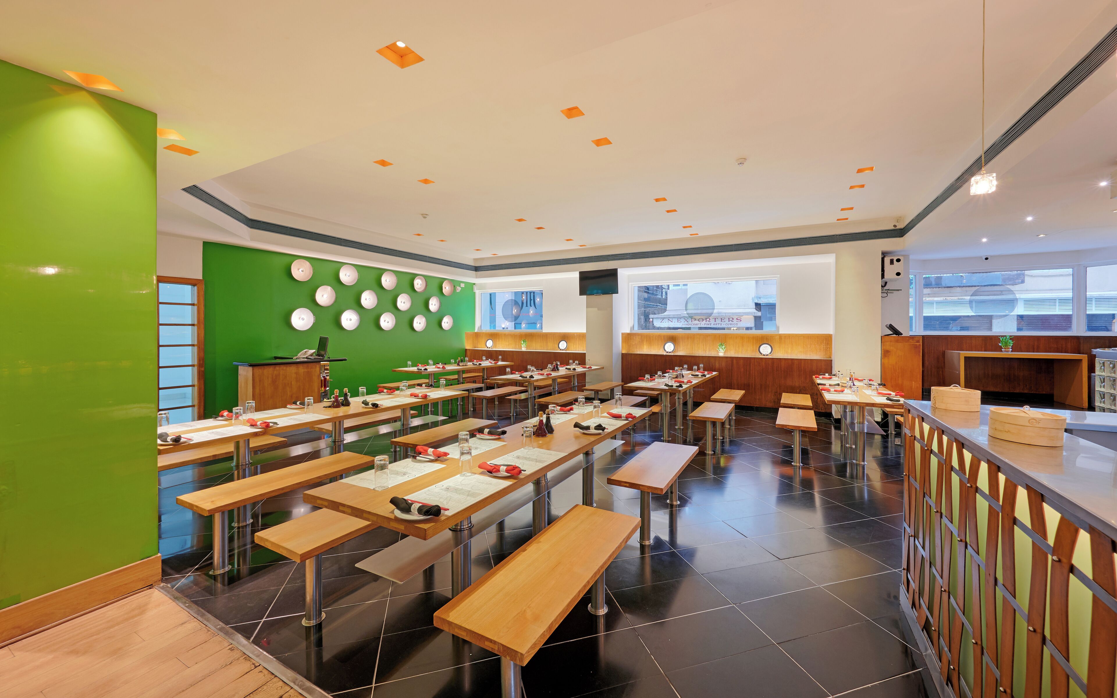 A modern restaurant with multiple tables and benches, a green accent wall, and a view of the city through large windows.