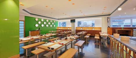A modern restaurant with multiple tables and benches, a green accent wall, and a view of the city through large windows.