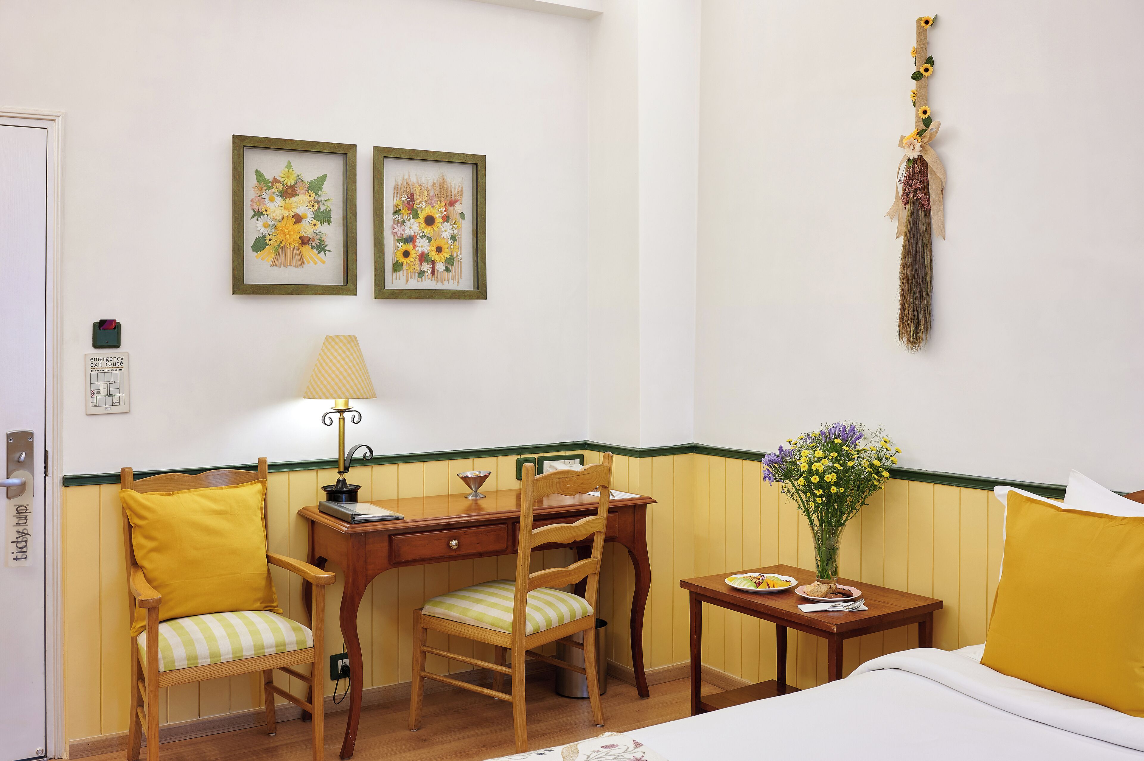 A hotel room with a bed, a desk with a chair, a small table with a vase of flowers, and two framed floral paintings on the wall.