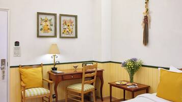 A hotel room with a bed, a desk with a chair, a small table with a vase of flowers, and two framed floral paintings on the wall.