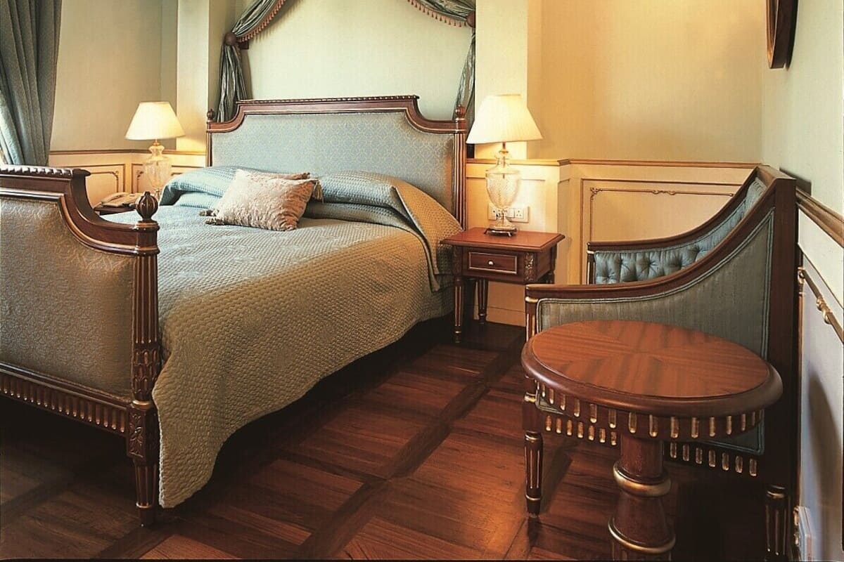A traditional bedroom with a four-poster bed, two bedside tables with lamps, a round wooden table, and a mirror on the wall.