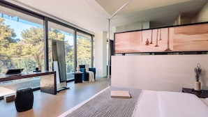 Une chambre d’hôtel moderne dotée d’une grande fenêtre, d’un lit, d’un bureau avec un ordinateur portable et d’un coin salon comprenant une chaise et une petite table.